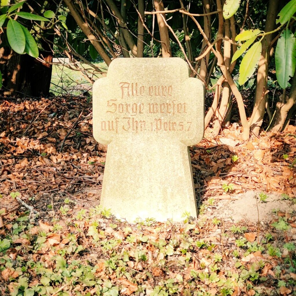 Ein Grabstein mit der Inschrift "Alle eure Sorge werfet auf Ihn. Petrus 5,7" steht zwischen Büschen und Laub, umgeben von frischem Grün.