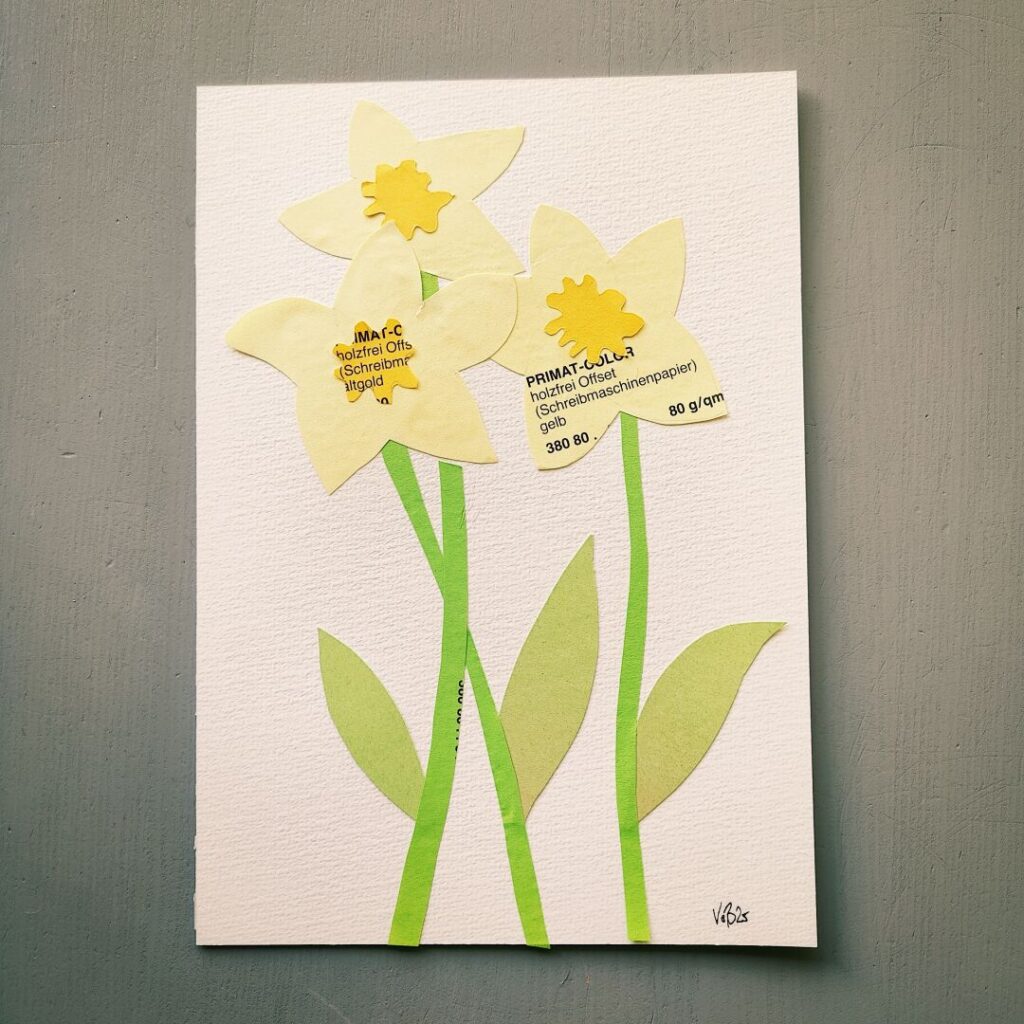 Zwei gelbe Papierblumen mit grünen Blättern auf einem weißen Hintergrund, versehen mit Etiketten, die Informationen über die verwendeten Materialien enthalten.