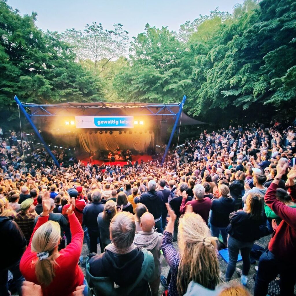Eine große Menschenmenge applaudiert begeistert bei einem Konzert in einer Freiluftbühne, umgeben von grünen Bäumen und stimmungsvollem Licht.