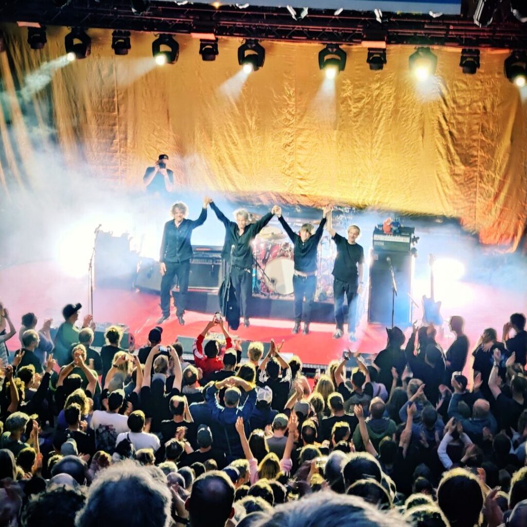 Eine Band verbeugt sich vor jubelndem Publikum auf einer Konzertbühne, umgeben von bunten Lichtern und Nebel, während die Musiker die Hände heben und die Zuschauer begeistert applaudieren.