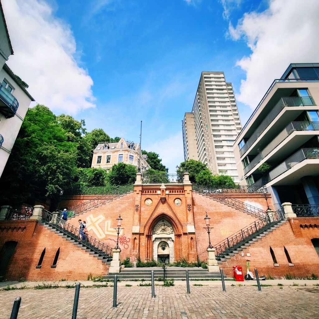Eine beeindruckende Treppenanlage aus rotem Ziegelstein führt zu einem historischen Gebäude, umgeben von modernen Wohnhäusern und üppigem Grün, unter einem strahlend blauen Himmel mit wenigen Wolken.
