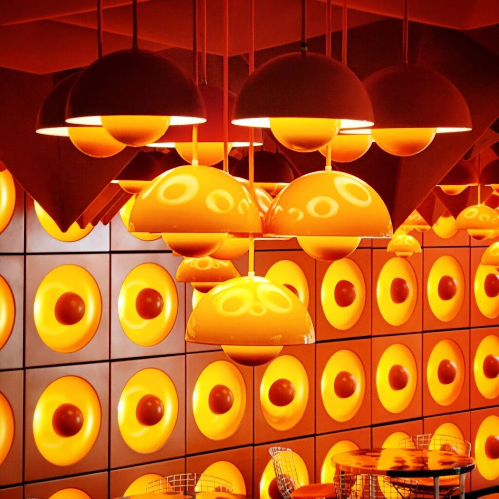 Leuchtende, orangefarbene Lampen hängen von der Decke in einem modernen Raum, während eine Wand mit runden, gelben Elementen in verschiedenen Größen dekoriert ist, die eine warme, einladende Atmosphäre schaffen.