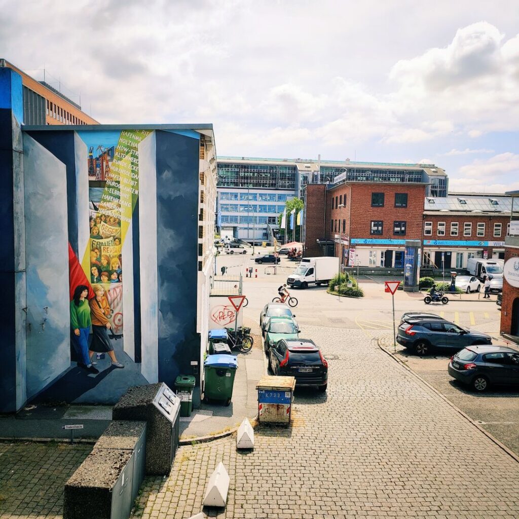 Eine lebendige Straßenszene zeigt ein Wandgemälde an einem Gebäude, während im Hintergrund moderne und historische Architektur sowie parkende Autos und Fahrräder zu sehen sind, unter einem klaren Himmel.