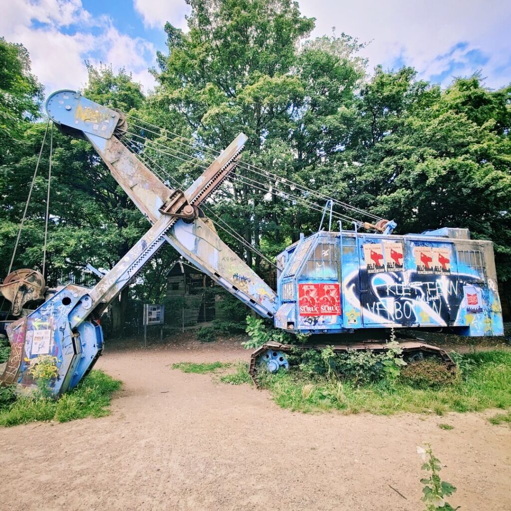 Eine verrostete, graffiti-bedeckte Baumaschine steht in einem grünen Umfeld, umgeben von Bäumen und Gras, und vermittelt einen Eindruck von Verfall und Urban Art.