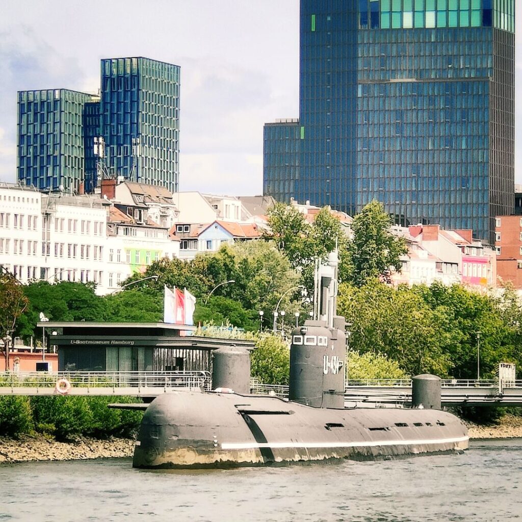 Ein U-Boot liegt am Ufer eines Flusses, umgeben von modernen Gebäuden und historischen Häusern, während im Hintergrund Bäume und eine Flagge sichtbar sind.