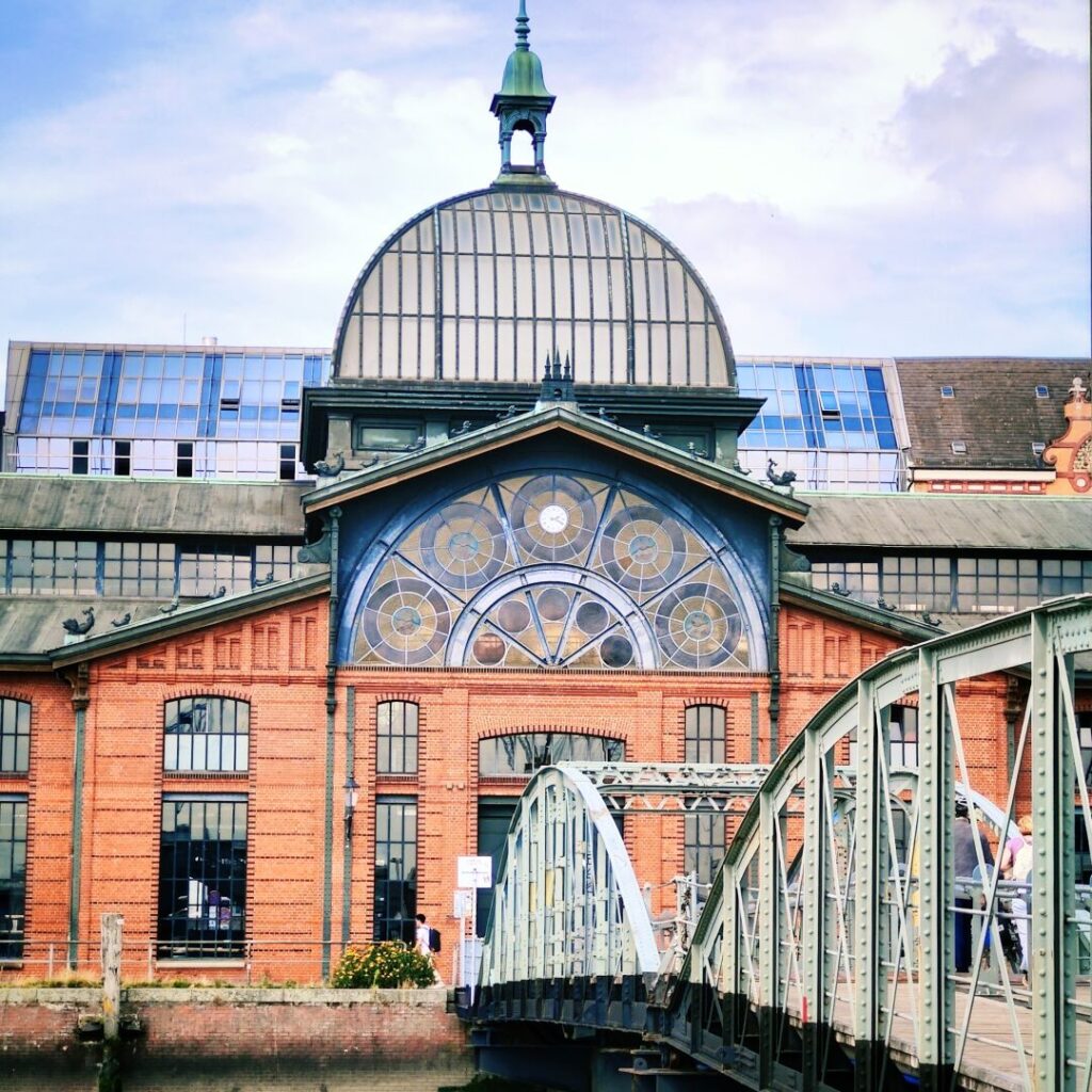 Historisches Gebäude mit einer markanten Kuppel und bunten Fenstern, umgeben von moderner Architektur, während eine Brücke im Vordergrund den Zugang zum Gebäude ermöglicht.