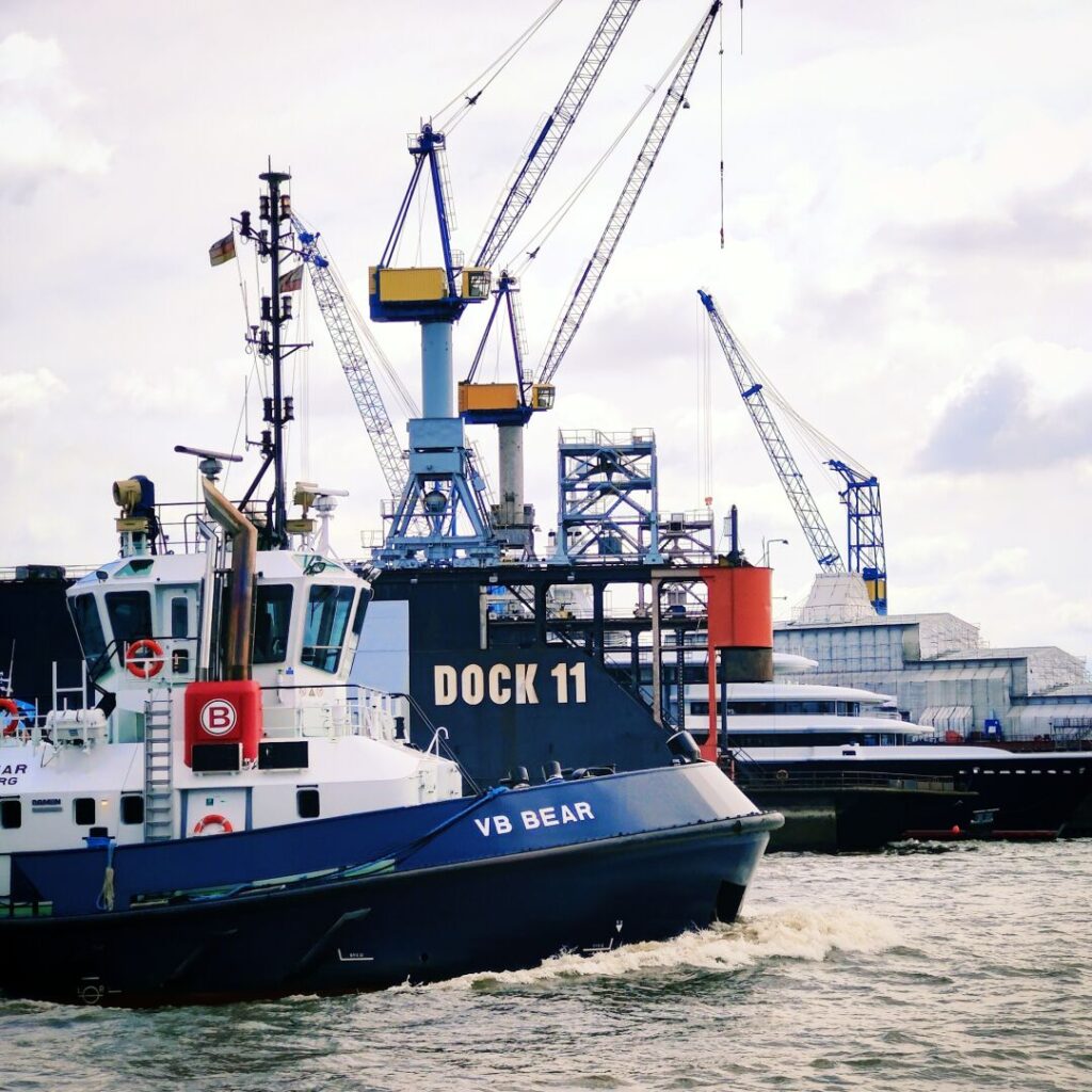 Ein Schlepper mit der Aufschrift "VB BEAR" fährt an einem Hafen mit Kränen und einem Dock mit der Bezeichnung "DOCK 11" vorbei, während sich im Hintergrund weitere Schiffe und Lagergebäude abzeichnen.