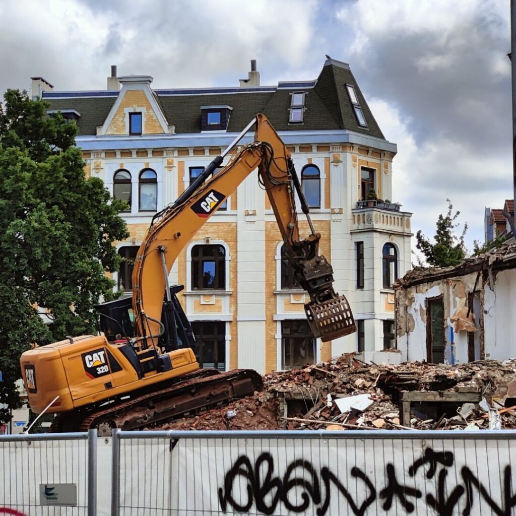 Ein gelber Bagger steht vor einem teilweise abgerissenen Gebäude, während im Hintergrund ein historisches Haus mit mehreren Fenstern und einem Dachfenster sichtbar ist, umgeben von einem Bauzaun und Graffiti.
