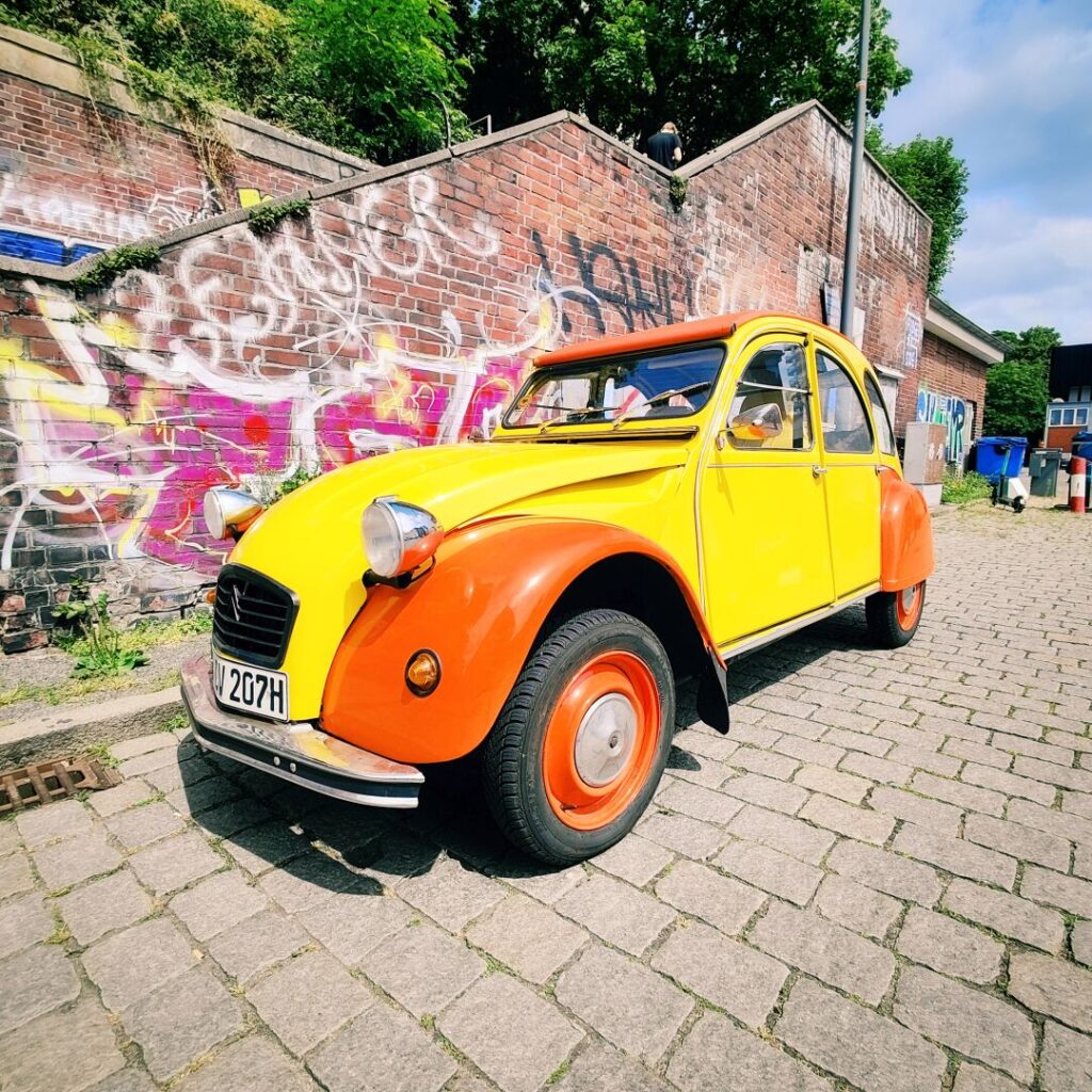 Ein bunt lackierter, gelb-orangefarbener Citroën 2CV steht auf einem gepflasterten Weg vor einer mit Graffiti verzierten Backsteinmauer, während im Hintergrund einige Personen zu sehen sind.