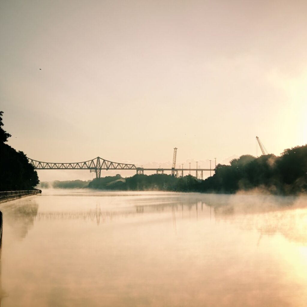 Ein ruhiger Fluss mit leichtem Nebel spiegelt die sanften Farben des Sonnenaufgangs wider, während eine Brücke und Baukräne im Hintergrund die Landschaft prägen.