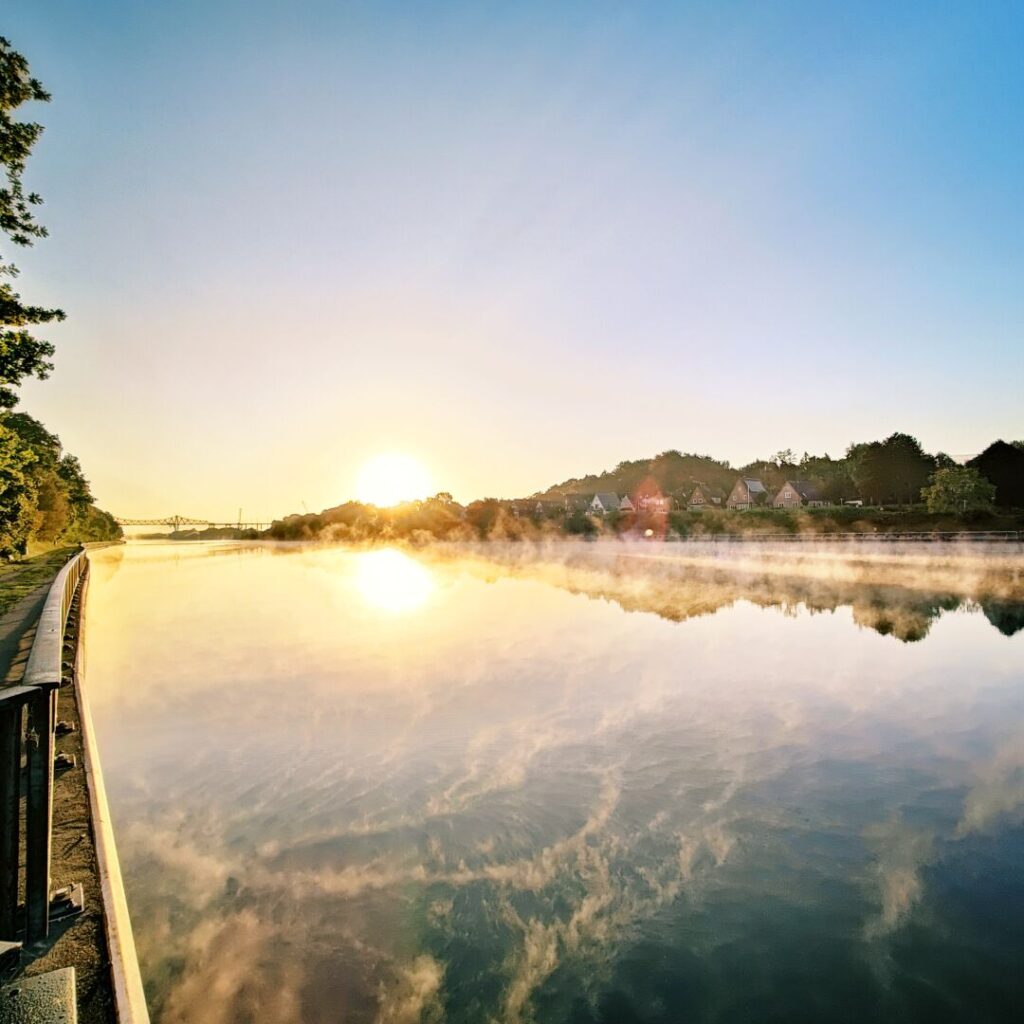 Ein ruhiger Fluss spiegelt die aufgehende Sonne wider, während der Nebel sanft über das Wasser zieht und die umgebenden Bäume und Häuser in warmes Licht taucht.