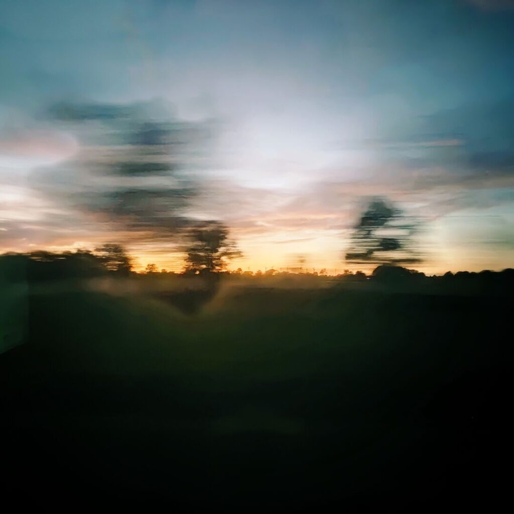 Ein verschwommener Sonnenuntergang mit bunten Wolken und Silhouetten von Bäumen, aufgenommen aus einem fahrenden Fahrzeug, vermittelt ein Gefühl von Bewegung und flüchtiger Schönheit.