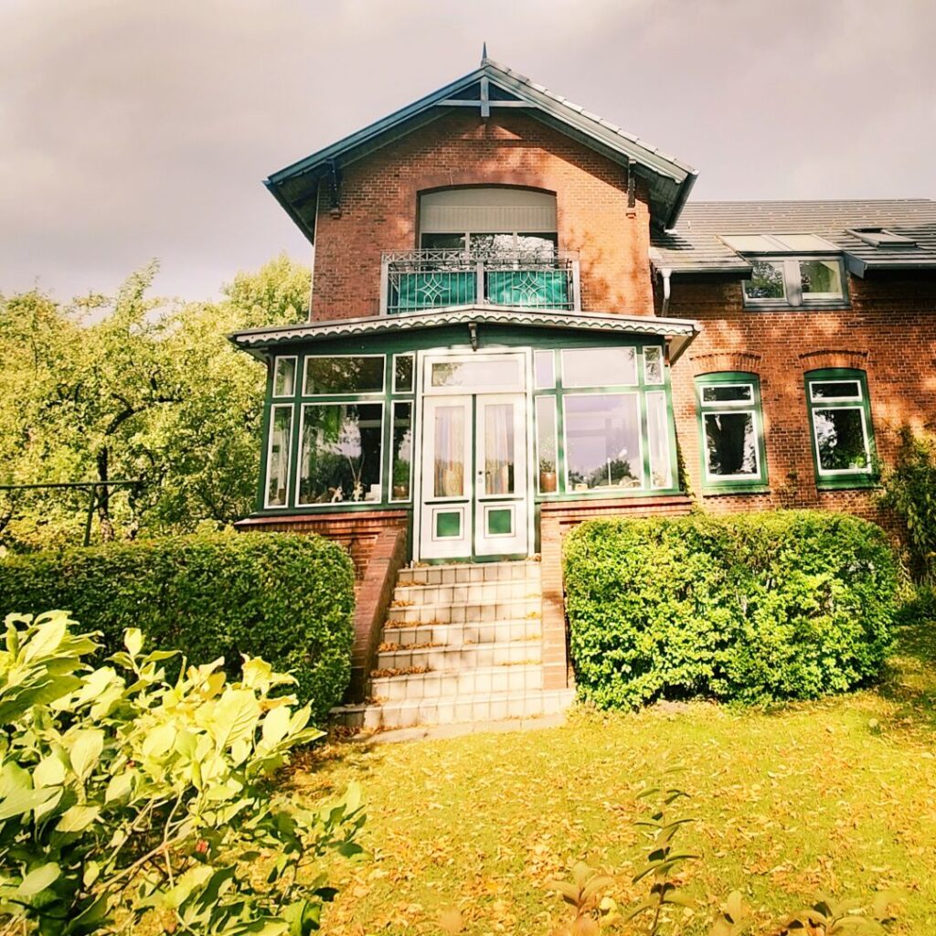 Ein rotes Backsteinhaus mit grünem Balkon und großen Fenstern, umgeben von gepflegten Hecken und einem herbstlichen Garten, unter einem bewölkten Himmel.