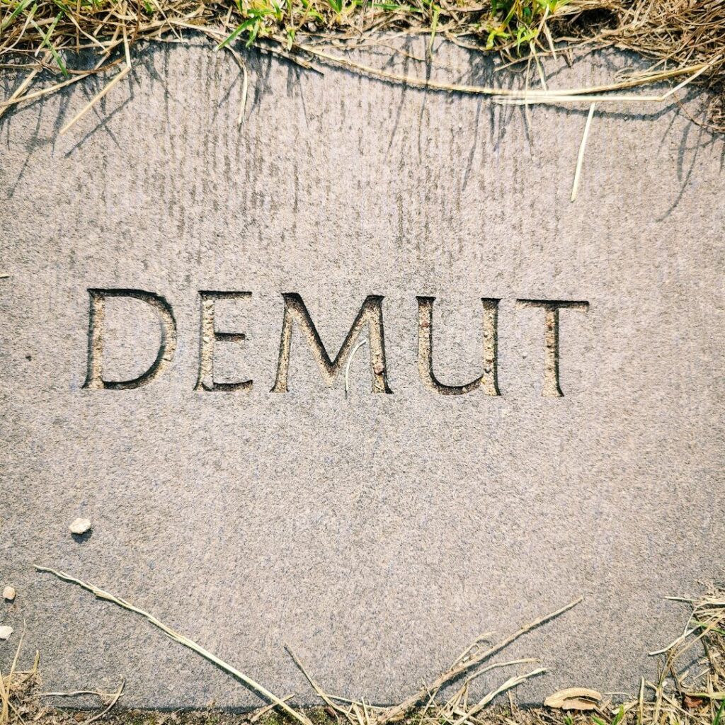 Eine gravierte Steinplatte mit dem Wort "DEMUT", umgeben von trockenem Gras und kleinen Steinen.