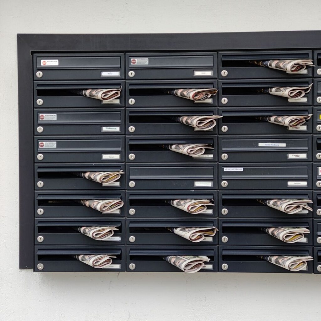 Zahlreiche Briefkästen mit eingeklemmten Zeitungen an einer Wand.