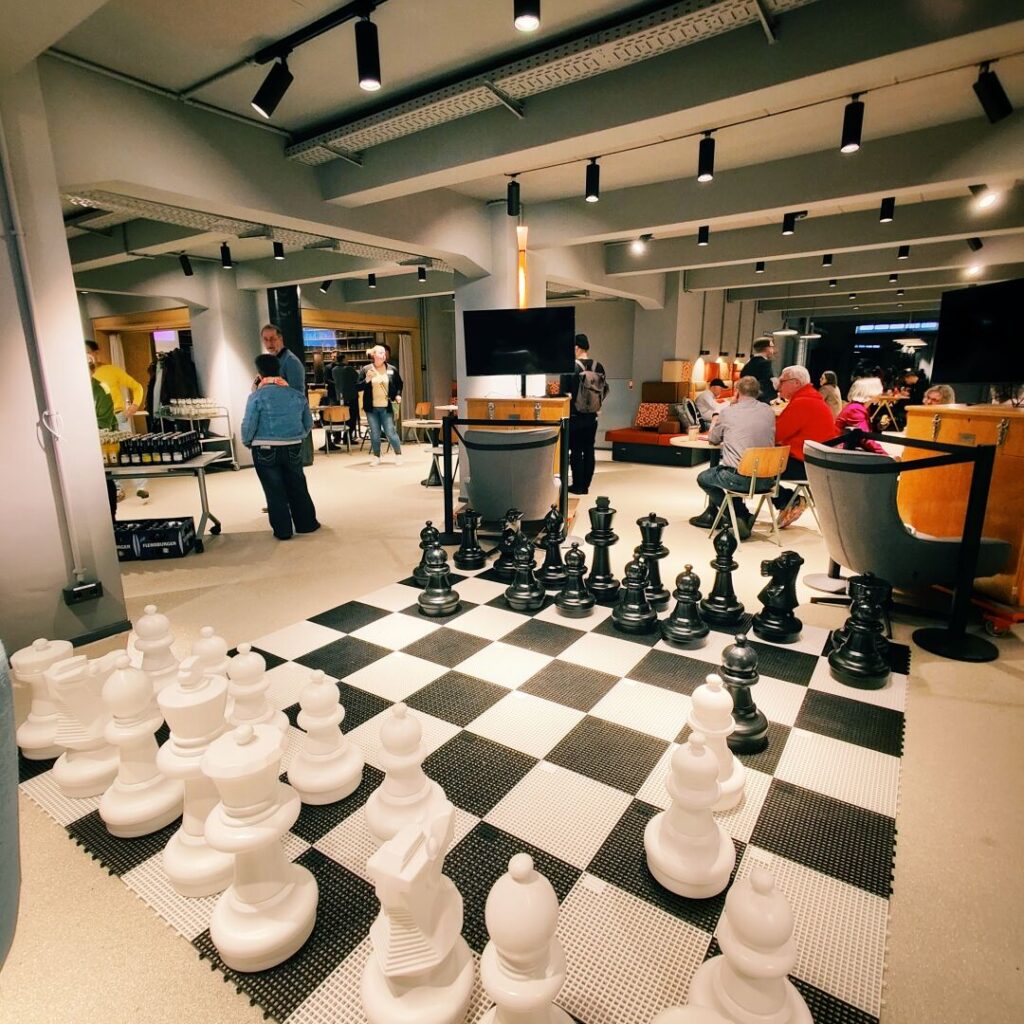 Eine große Schachbrettinstallation mit weißen und schwarzen Figuren in einem modernen Veranstaltungsraum, umgeben von Gästen.