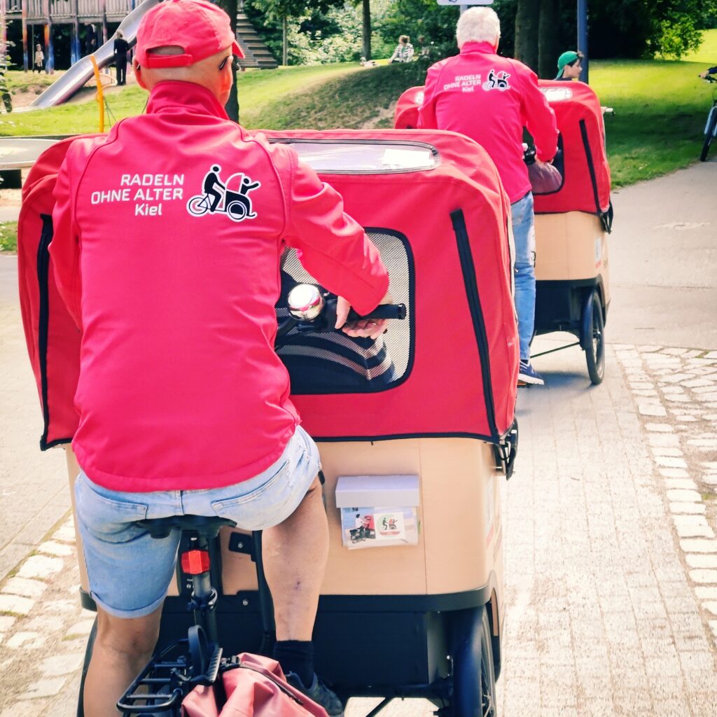 Drei Personen in roten Jacken fahren auf Fahrradrikschas durch einen Park in Kiel.