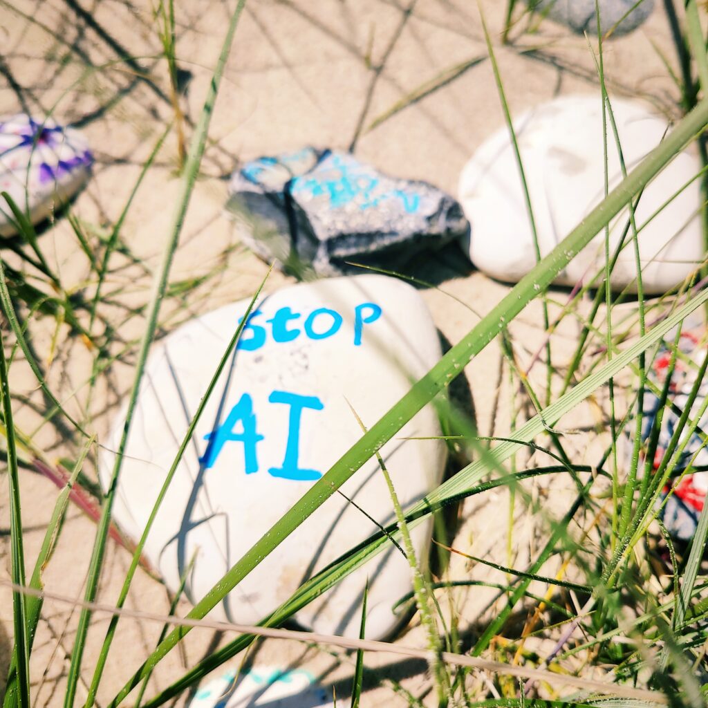 Weißer Stein mit der Aufschrift "stop AI" liegt im Gras auf sandigem Boden.
