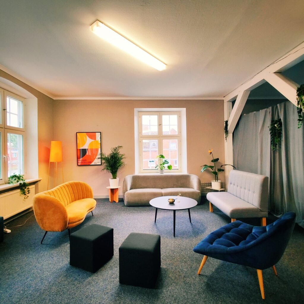 Gemütlicher Raum mit modernen Sitzmöbeln in verschiedenen Farben, einem runden Tisch in der Mitte, Pflanzen und einem großen Fenster, das Tageslicht hereinlässt, sowie einem bunten Kunstwerk an der Wand.