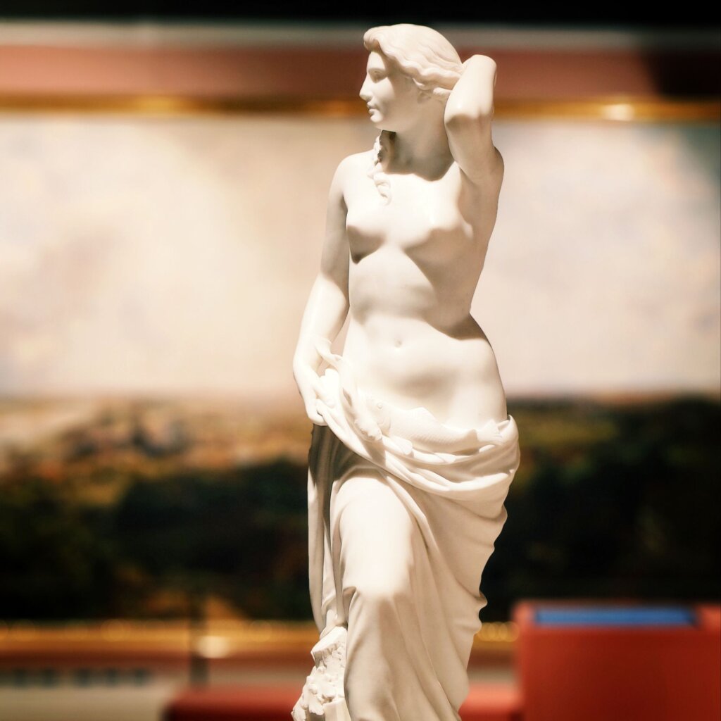 Eine elegante, weiße Marmorstatue einer weiblichen Figur mit langen Haaren, die einen Stoff um ihre Hüften drapiert, steht vor einem unscharfen Hintergrund mit Landschaftsmalerei.