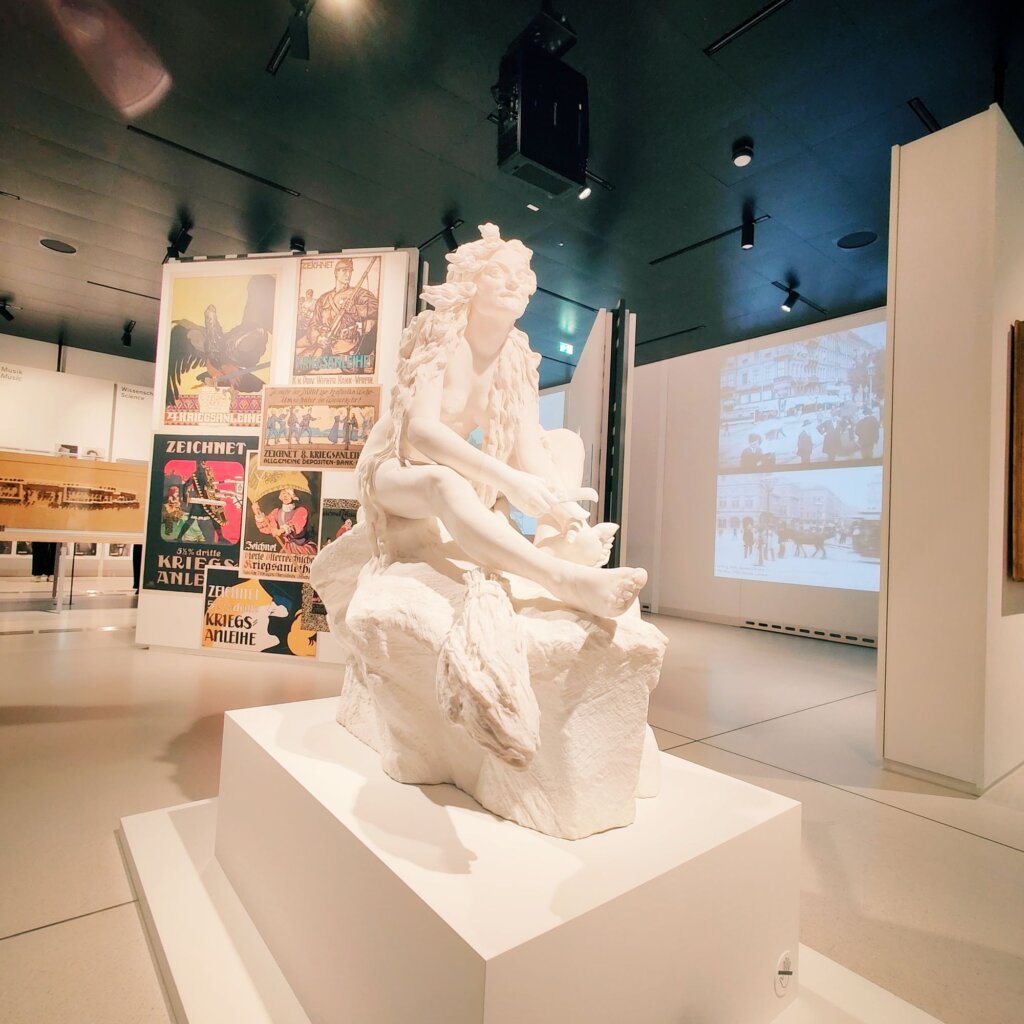 Eine weiße Skulptur einer sitzenden Figur mit langen Haaren, die auf einem Podest in einem modernen Ausstellungsraum präsentiert wird, während im Hintergrund historische Plakate und Schwarz-Weiß-Fotos an den Wänden zu sehen sind.