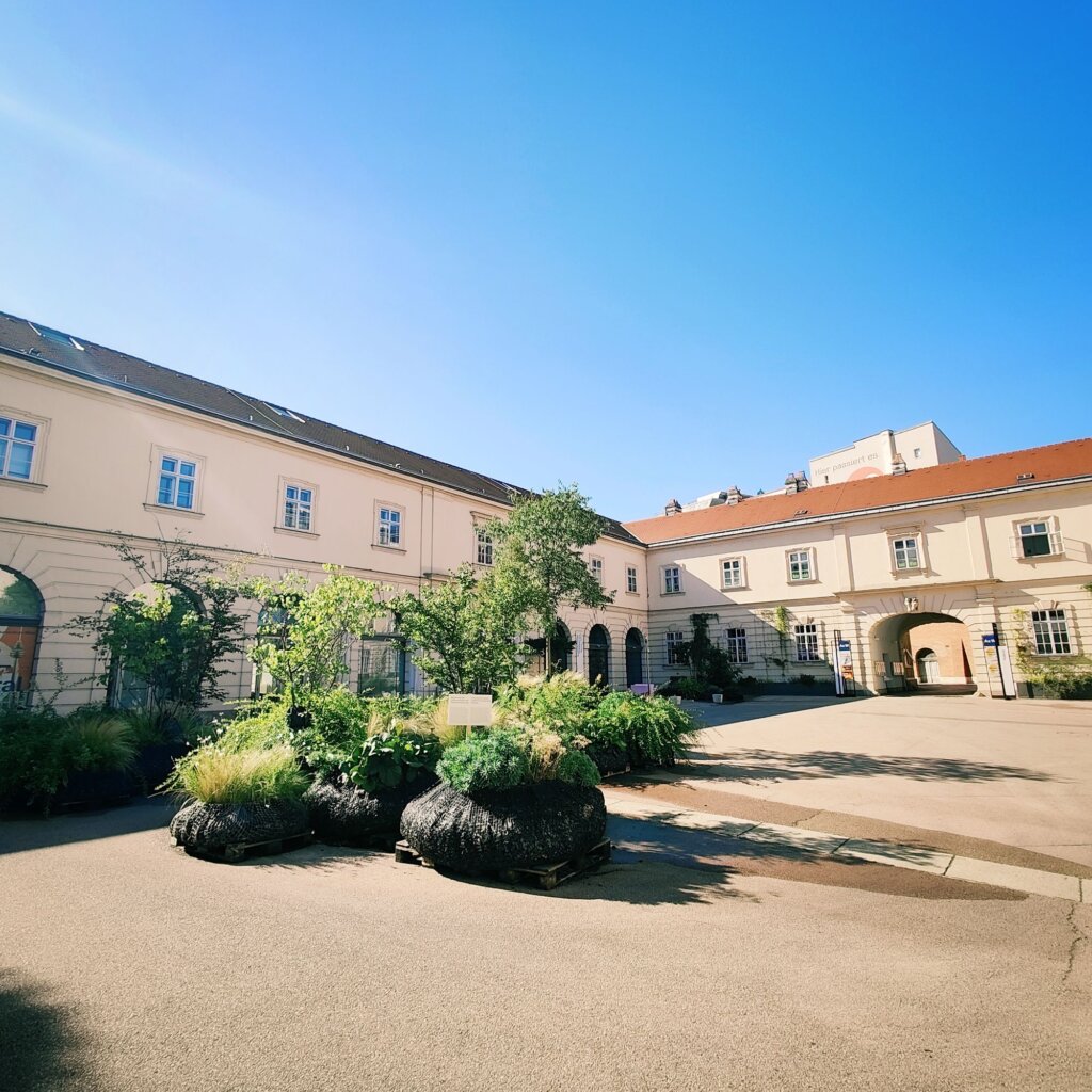 Eine sonnige Innenhofansicht mit historischen Gebäuden, umgeben von üppigem Grün und Pflanzen in großen Töpfen, unter einem klaren blauen Himmel.