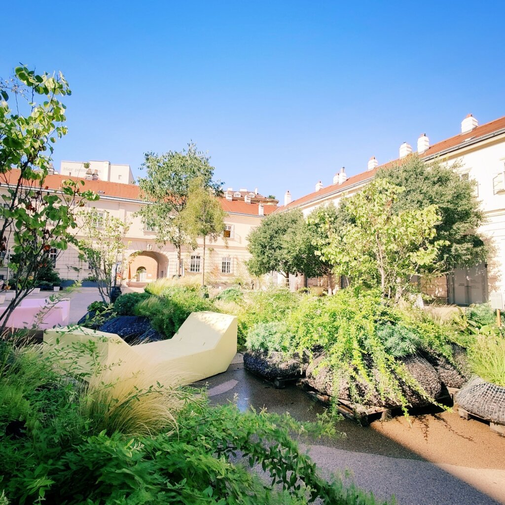 Ein sonniger Innenhof mit üppigem Grün, verschiedenen Bäumen und Sträuchern, einem kleinen Wasserlauf und einer gelben Sitzgelegenheit, umgeben von historischen Gebäuden mit roten Dächern und klarem blauen Himmel.
