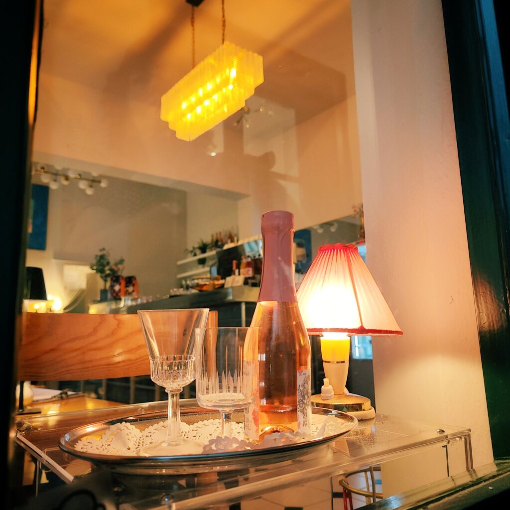 Ein elegantes Tablett mit einer Flasche Roséwein und zwei Gläsern steht auf einem Tisch, während eine kleine Lampe mit rotem Schirm und eine stilvolle Deckenlampe eine gemütliche Atmosphäre im Hintergrund schaffen.
