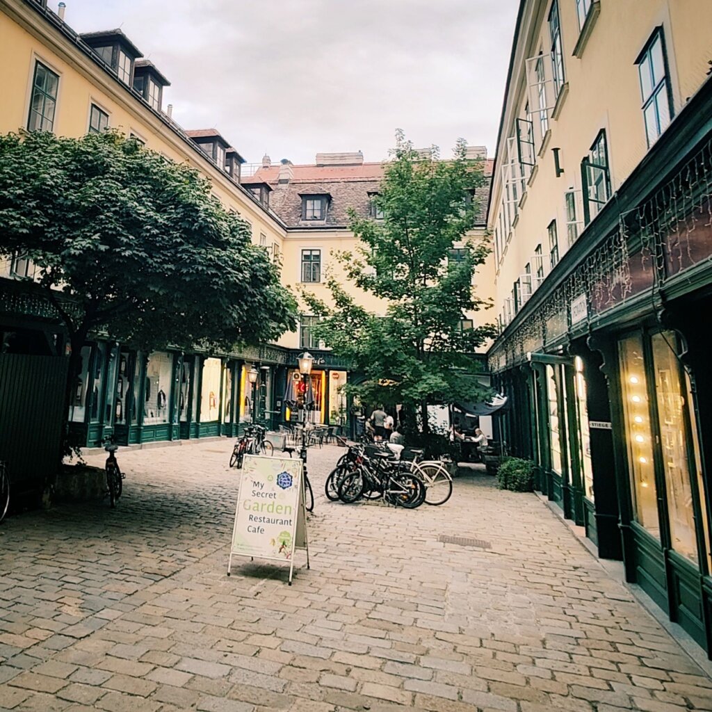 Eine malerische Gasse mit historischen Gebäuden, Bäumen und Fahrrädern, in der ein Schild für ein Restaurant-Café namens "My Secret Garden" steht, während sich im Hintergrund Menschen unterhalten.