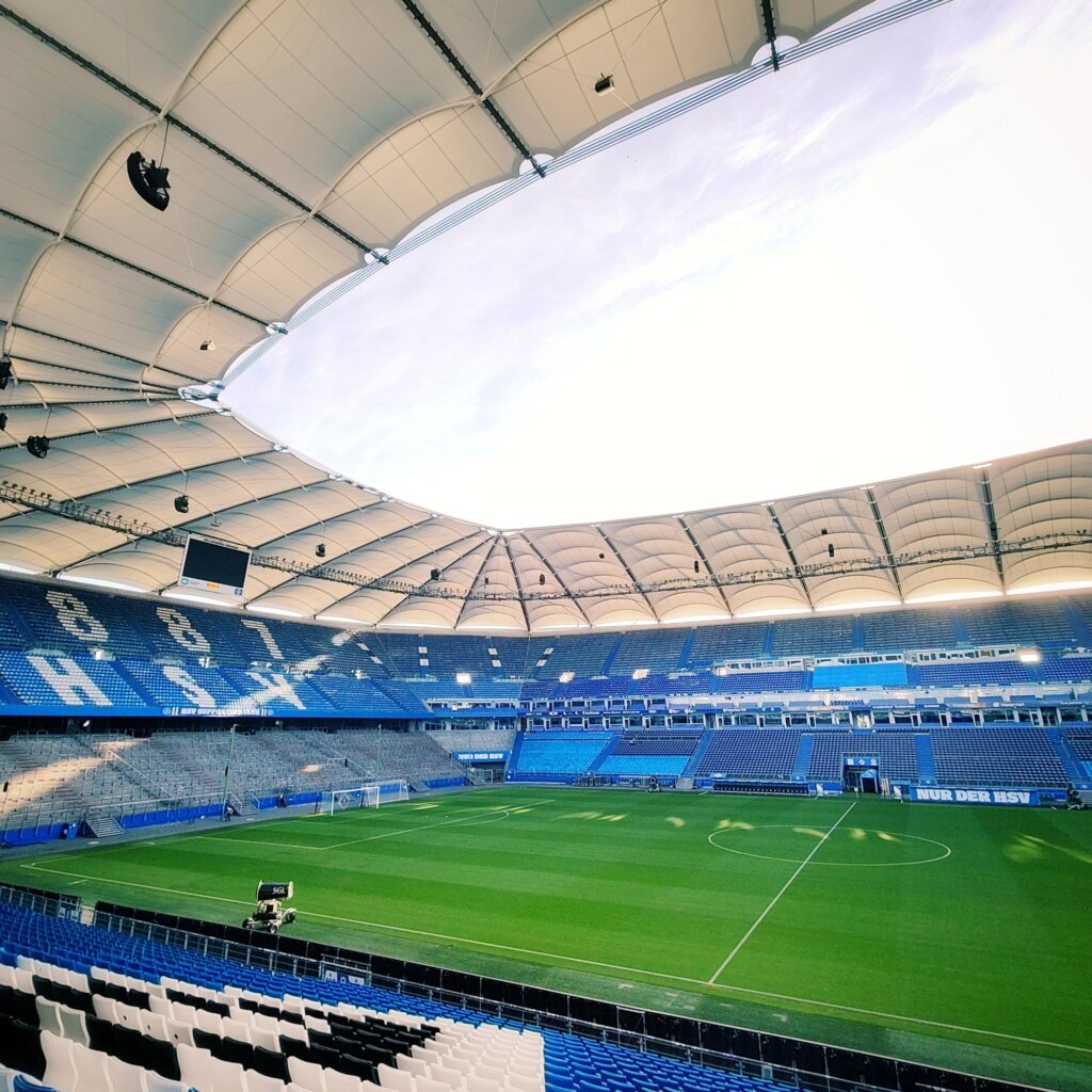 Das Bild zeigt das Innere eines modernen Fußballstadions mit einem großen, offenen Dach und einer großzügigen Sitzplatzanordnung. Die Tribünen sind in verschiedenen Blautönen gestaltet, während das Spielfeld gut gepflegt und mit Markierungen versehen ist. Im Hintergrund sind große LED-Bildschirme und Lautsprecher sichtbar. Die Atmosphäre wirkt einladend und bereit für ein spannendes Spiel. Der Himmel ist klar, was auf einen sonnigen Tag hindeutet. Die gesamte Szenerie vermittelt ein Gefühl von Sportbegeisterung und Gemeinschaft.