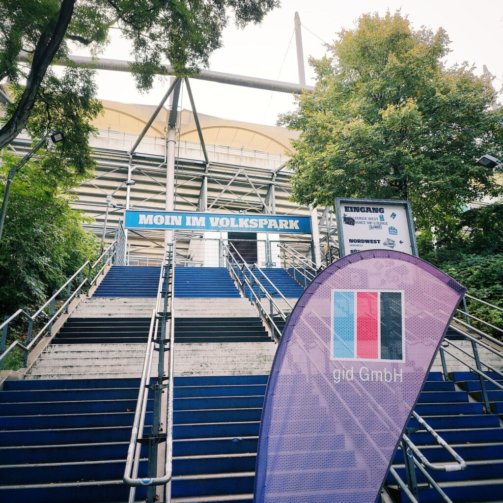 Treppenaufgang zum Volksparkstadion mit einem großen blauen Schild, das "Moin im Volkspark" zeigt. Auf der linken Seite steht ein Werbebanner der Firma gid GmbH in lila und pink. Die Treppen sind in verschiedenen Blautönen gestrichen und von Bäumen umgeben. Im Hintergrund sind die charakteristischen Strukturen des Stadions sichtbar, die durch große Stahlträger und ein gewölbtes Dach geprägt sind. Der Eingang ist gut beleuchtet und einladend gestaltet, was auf bevorstehende Veranstaltungen hinweist.