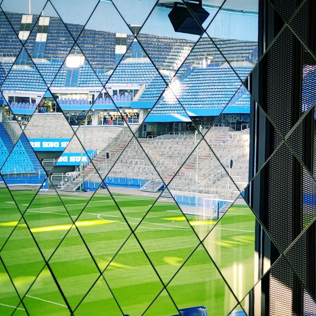 Blick auf ein Fußballstadion durch ein Gitterfenster. Im Vordergrund ist der gepflegte Rasen des Spielfelds zu sehen, mit klaren Linien und einem Tor im Hintergrund. Die Tribünen sind teilweise besetzt, mit blauen Sitzen, die eine moderne Atmosphäre schaffen. Im oberen Bereich des Bildes sind weitere Zuschauer und Einrichtungen sichtbar. Das Licht fällt durch das Fenster und erzeugt eine lebendige Stimmung im Stadion. Die Architektur ist zeitgemäß und spiegelt die Dynamik des Fußballs wider.