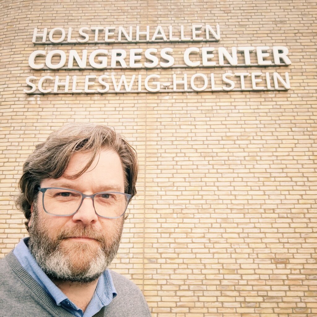 Ein Mann mit Brille und Bart steht vor der Fassade des Holstenhallen Congress Centers in Schleswig-Holstein. Die Wand ist aus hellen Ziegeln und trägt den Schriftzug "HOLSTENHALLEN CONGRESS CENTER SCHLESWIG-HOLSTEIN" in großen, modernen Buchstaben. Der Mann trägt einen grauen Pullover über einem blauen Hemd und blickt direkt in die Kamera. Die Umgebung wirkt freundlich und einladend, was auf eine Veranstaltung oder ein Treffen in diesem Kongresszentrum hindeutet.