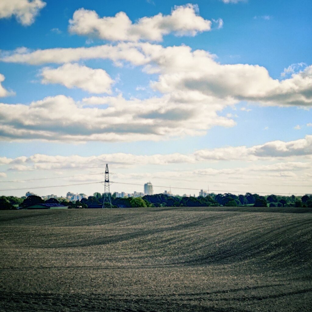Weite, braune Felder erstrecken sich im Vordergrund, während im Hintergrund eine Stadtlandschaft mit modernen Gebäuden sichtbar ist. Der Himmel ist blau mit vielen weißen, flauschigen Wolken. Eine Stromleitung verläuft diagonal durch das Bild und verbindet die ländliche Umgebung mit der urbanen Kulisse. Die Szenerie vermittelt einen Kontrast zwischen Natur und Stadtleben, wobei die Felder gut bearbeitet und die Bäume am Rand der Stadt grün leuchten.