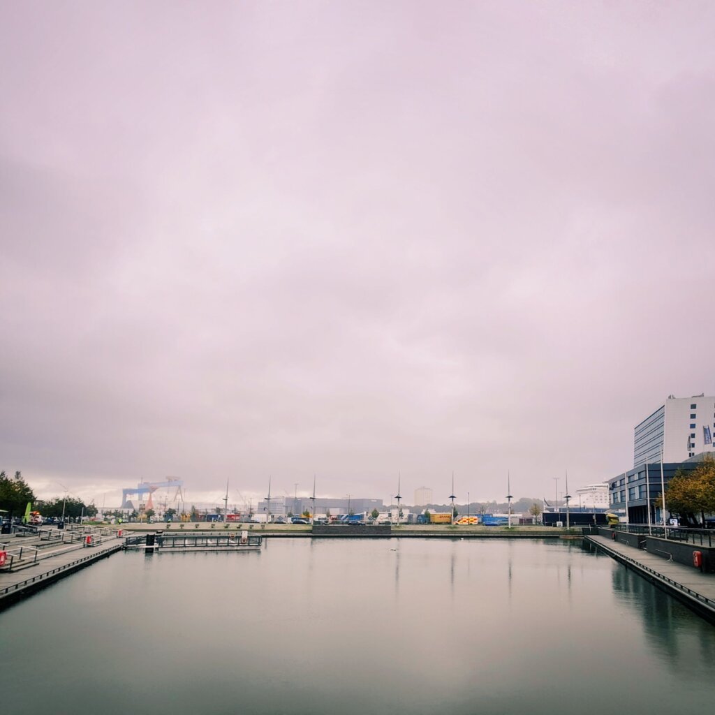 Eine ruhige Hafenansicht an einem bewölkten Tag. Der Wasserbereich spiegelt die grauen Wolken wider, während am Ufer moderne Gebäude und eine Promenade sichtbar sind. Auf der linken Seite sind Holzstege und einige Laternen zu erkennen. Im Hintergrund sind verschiedene Schiffe und Container zu sehen, die auf eine geschäftige Hafenaktivität hinweisen. Die Atmosphäre wirkt ruhig und leicht melancholisch, geprägt von den sanften Farben des Himmels und der Umgebung.