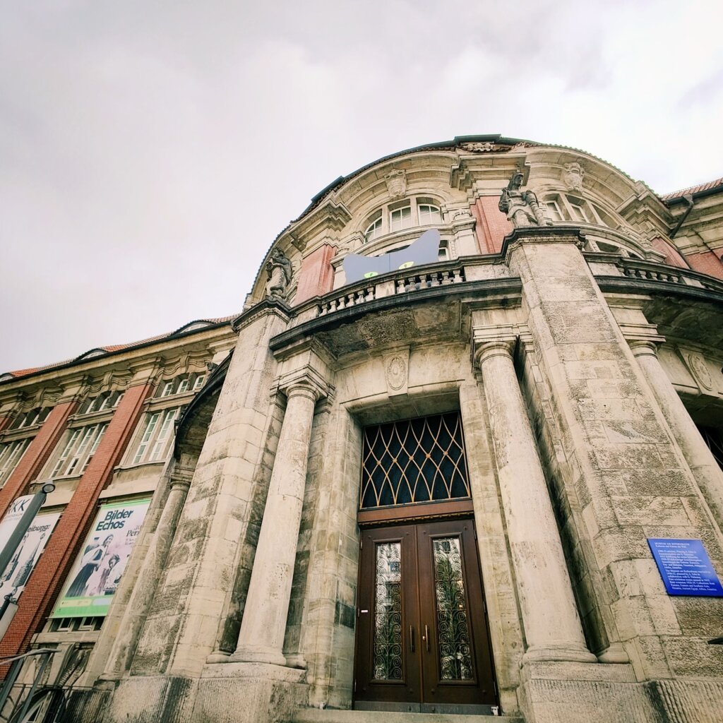 Historisches Gebäude mit beeindruckender Architektur, das von einer Vielzahl von Säulen und einem geschwungenen Dach geprägt ist. Die Fassade zeigt eine Kombination aus grauem Stein und roten Ziegeln. Über dem Eingang befinden sich kunstvolle Fenster mit einem dekorativen Gitter. An den Seiten sind Plakate angebracht, die auf aktuelle Ausstellungen hinweisen. Der Himmel ist bewölkt, was dem Bild eine melancholische Stimmung verleiht. Die Perspektive ist von unten nach oben gerichtet, wodurch die Höhe des Gebäudes betont wird.
