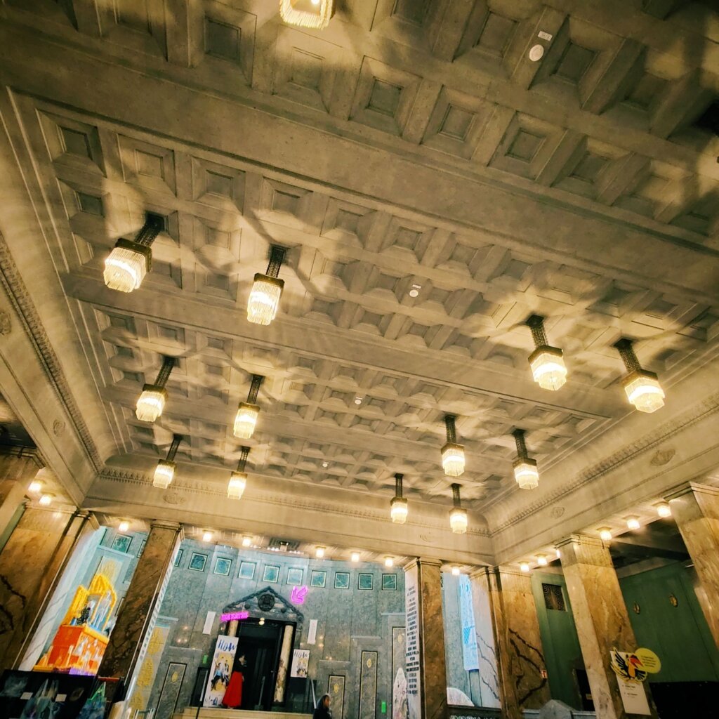 Der Blick auf die Decke eines großzügigen Innenraums zeigt ein kunstvolles, geometrisches Stuckdesign mit quadratischen und rechteckigen Mustern. An der Decke hängen mehrere elegante, gläserne Pendelleuchten, die warmes Licht spenden und interessante Schattenmuster erzeugen. Die Wände sind mit marmorähnlichem Material verkleidet, das dem Raum eine edle Atmosphäre verleiht. Im Hintergrund sind farbige Plakate und Kunstwerke zu sehen, die dem Raum einen modernen Touch verleihen. Der Eingang ist durch eine große, geschwungene Tür gekennzeichnet.