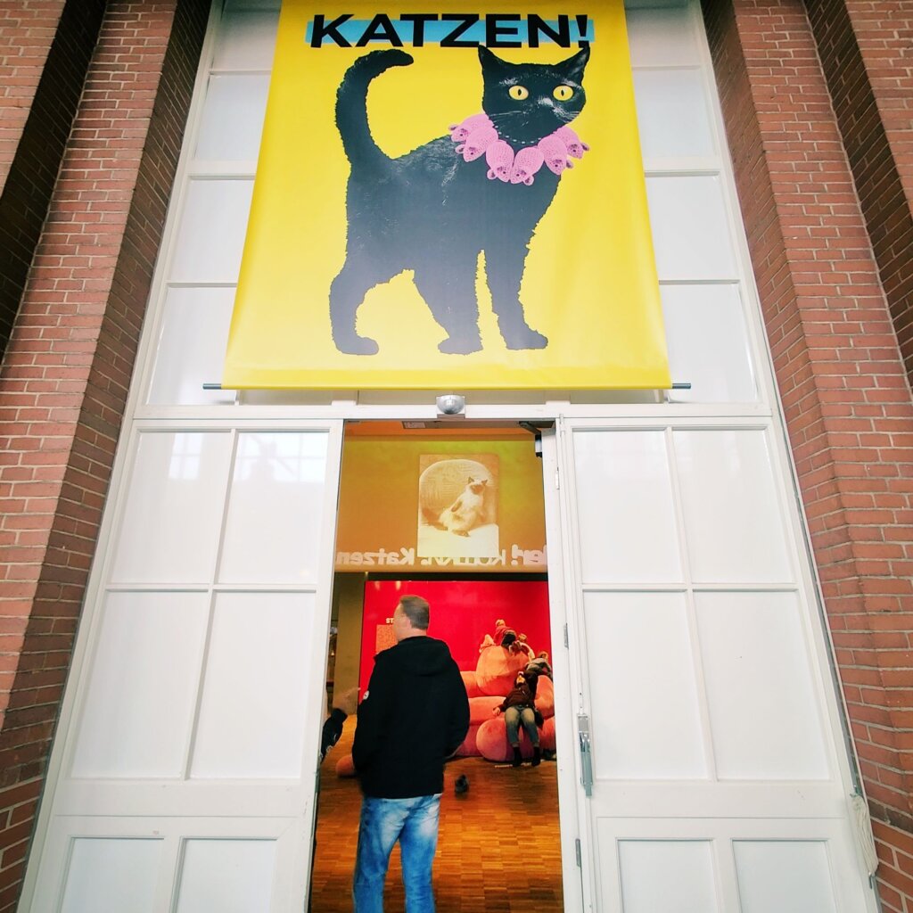 Ein großer, gelber Banner mit der Aufschrift "KATZEN!" zeigt eine schwarze Katze mit einem pinken Kragen. Der Eingang zu einer Ausstellung ist sichtbar, durch die eine Person in schwarzer Jacke und blauen Jeans geht. Im Hintergrund sind verschwommene Silhouetten von Besuchern und große, runde, orangefarbene Objekte zu erkennen, die möglicherweise Sitzgelegenheiten sind. Die Wände des Ausstellungsraums sind in warmen Farben gehalten, was eine einladende Atmosphäre schafft.