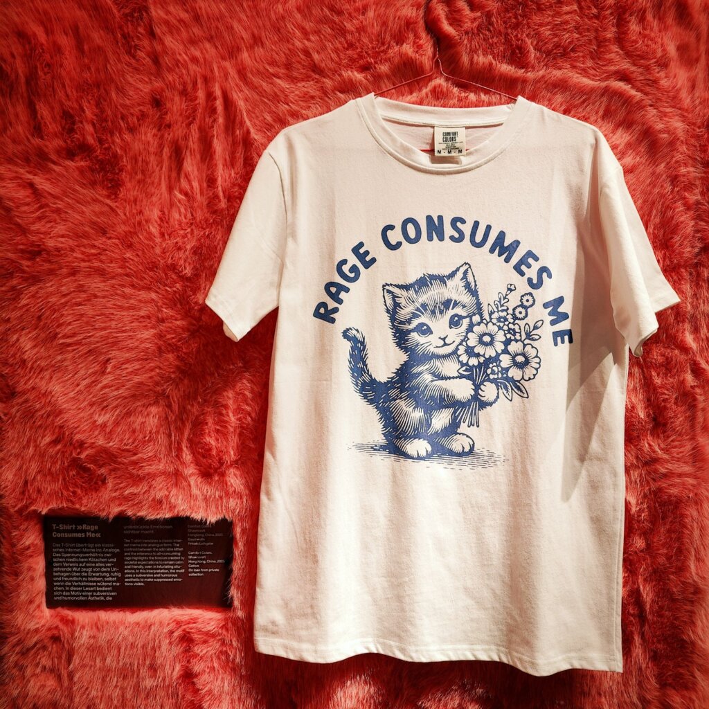 Ein weißes T-Shirt hängt vor einem roten, flauschigen Hintergrund. Auf dem Shirt ist eine Illustration einer niedlichen Katze zu sehen, die mit einer Hand Blumen hält. Über der Katze steht in blauer Schrift der Spruch "RAGE CONSUMES ME". Das Design kombiniert humorvolle Elemente mit einem verspielten, künstlerischen Stil. Neben dem Shirt befindet sich ein schwarzes Schild mit Informationen über das T-Shirt und dessen Bedeutung. Die gesamte Komposition vermittelt eine lebendige und kreative Atmosphäre.