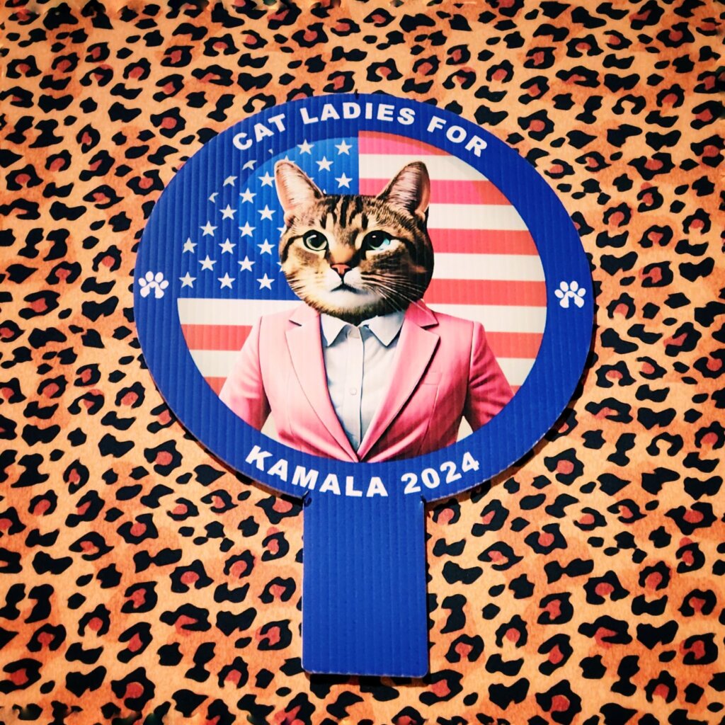Ein kreatives Plakat zeigt eine Katze mit menschlichem Oberkörper, die einen rosa Anzug trägt. Der Hintergrund ist mit einer amerikanischen Flagge gestaltet, die teilweise sichtbar ist. Oben steht der Schriftzug "CAT LADIES FOR", während unten "KAMALA 2024" zu lesen ist. Das gesamte Motiv ist auf einem leopardenmusterartigen Untergrund platziert, was dem Bild einen auffälligen und humorvollen Charakter verleiht. Die Kombination aus politischen und tierischen Elementen spricht eine spezielle Zielgruppe an.