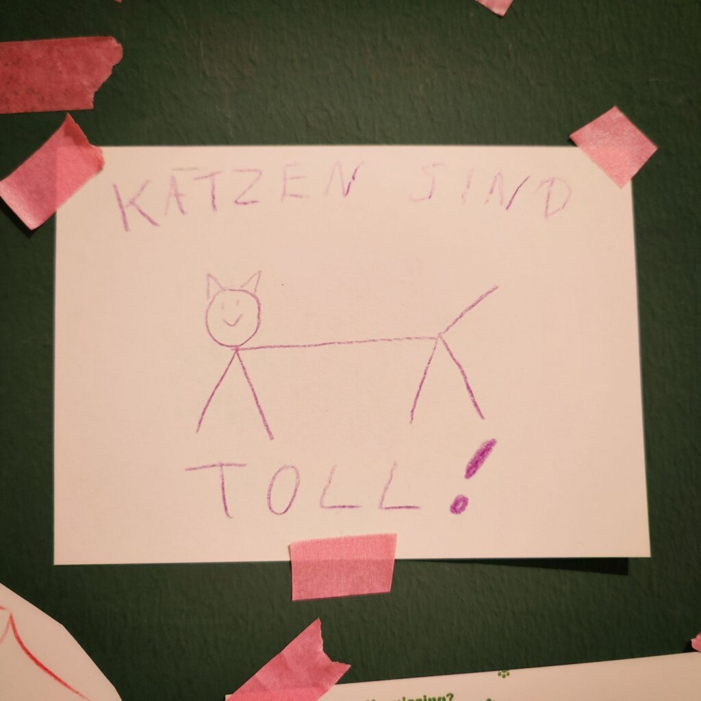 Auf einem weißen Blatt Papier, das an einer grünen Wand befestigt ist, steht in lila Schrift der Text "KATZEN SIND TOLL!" über einer einfachen Zeichnung einer Katze. Die Katze ist mit einfachen Linien dargestellt, hat einen runden Kopf mit einem lächelnden Gesicht und spitzen Ohren. Der Körper ist lang und schlank, mit vier Beinen. Das Blatt ist mit pinkem Klebeband an den Ecken befestigt, was dem Bild eine kindliche und kreative Note verleiht.